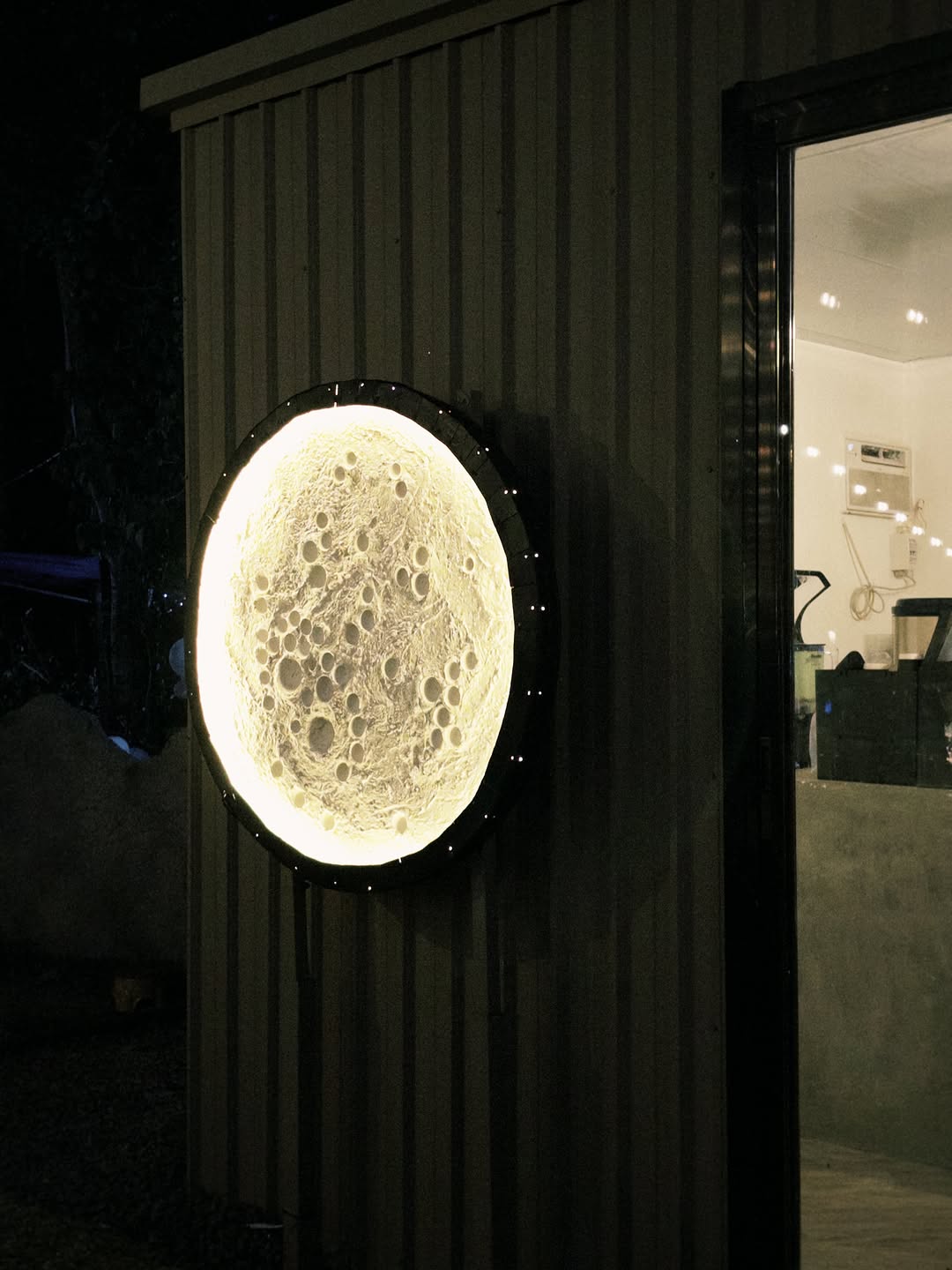 Moon Coffee 2.0 moon lamp on the side wall at night, bright interior espresso bar visible through the door