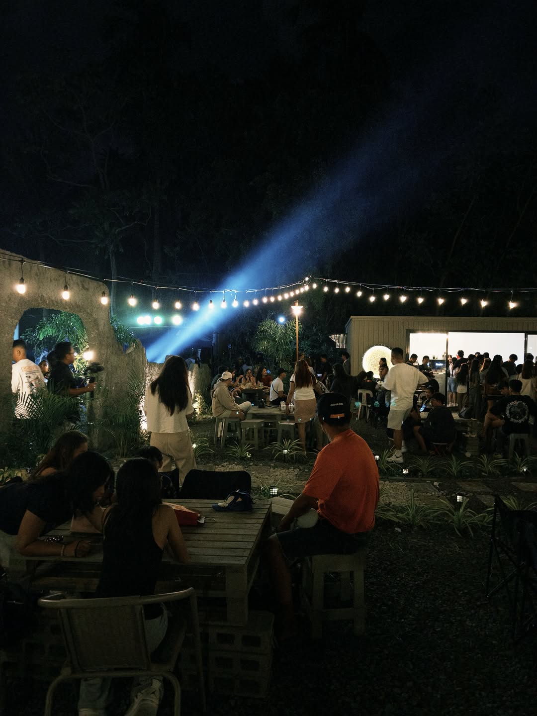 Blue beam spotlight over Moon Coffee outdoor area — string lights, stone walls, and crowded tables at night